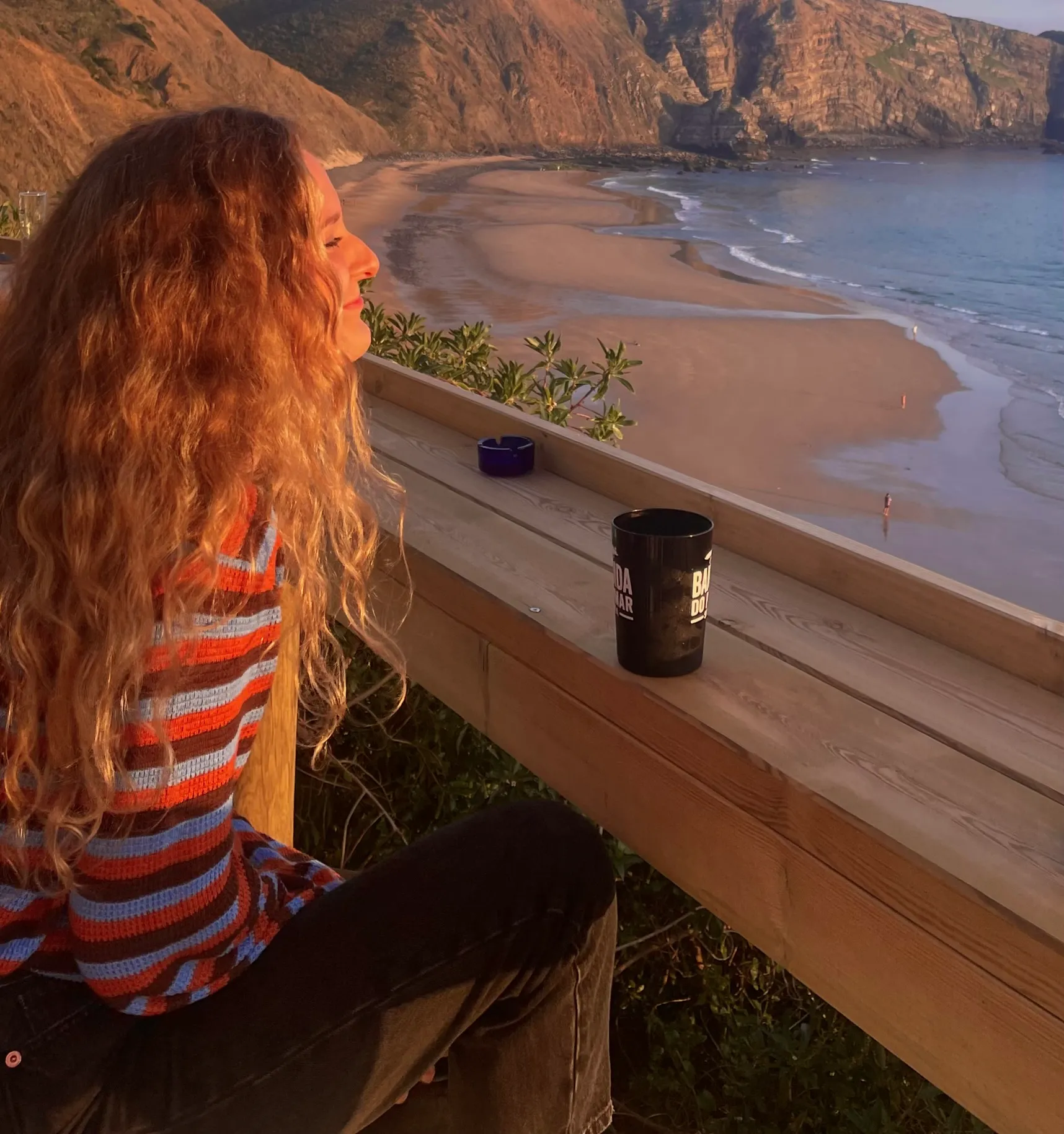 Eleonora sitting on a clifftop terrace at sunset, looking out over a beach